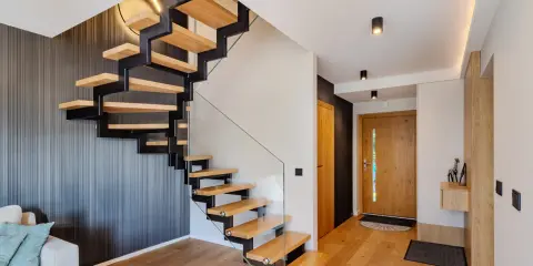 Nowoczesne schody zabiegowe z drewna i metalu, z szklaną balustradą, prowadzące do holu z drewnianymi drzwiami.