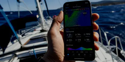 Sur un voilier, une main tient un smartphone affichant une application météo marine gratuite avec les prévisions pour Ibiza.
