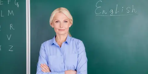 Nauczycielka angielskiego z uśmiechem, stojąca przed tablicą z napisem "English". Marzy o wcześniejszej emeryturze dla nauczycieli.
