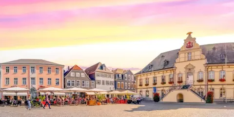 Ein Marktplatz mit Cafés und historischen Gebäuden, der an das "Venedig Westfalens" erinnert, unter einem farbenfrohen Himmel.