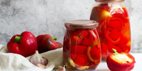 Frische rote Paprika, Knoblauch und Pfefferkörner neben zwei Gläsern mit eingelegter Paprika. Perfekt zum Paprika einkochen mit Weck-Gläsern.