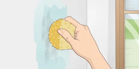 Mano limpiando una mancha en una pared blanca con una esponja. Aprende como limpiar paredes blancas fácilmente.