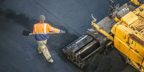 Arbeiter schaufelt heißes Bitumen auf eine Straße, während ein Asphaltfertiger das Material verteilt.