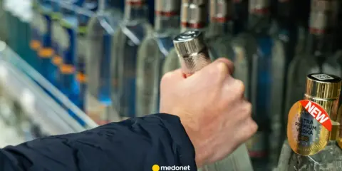 Dłoń sięga po butelkę wódki, a artykuł na medonet.pl analizuje wpływ alkoholu, w tym piwa, na białe krwinki.