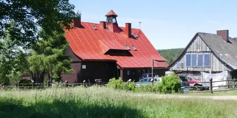 Chatka Górzystów z czerwonym dachem i drewnianymi budynkami. W tle las, a przed budynkami zielona łąka.