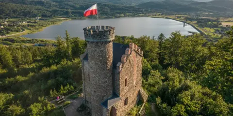 Kamienna wieża z polską flagą na tle jeziora i gór. Wspaniałe **cieplice atrakcje** dla miłośników historii i przyrody.