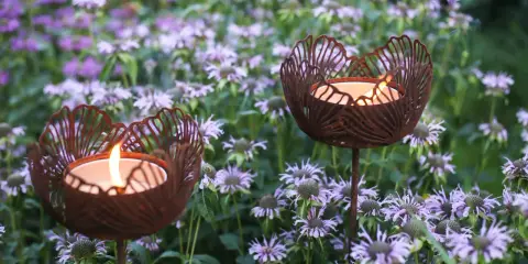 Två rostiga ljushållare i form av blommor lyser upp en rabatt med lila blommor.