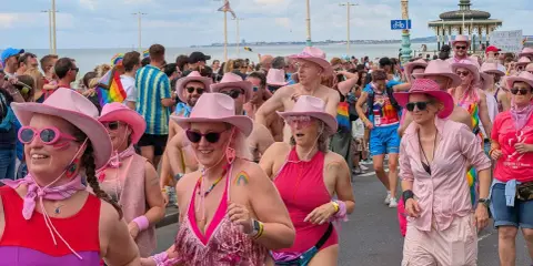 A vibrant Brighton gay pride parade, with many participants wearing pink cowboy hats and rainbow-themed attire, celebrating joyfully by the sea.