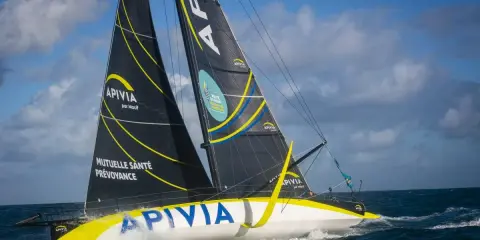 Le voilier Apivia, skippé par Alex Thomson, fend les vagues sous un ciel bleu et nuageux.