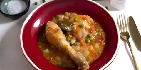 Un delicioso pollo a la jardinera en un cuenco rojo, con guisantes, zanahoria y tomate. Acompañado de sal, laurel y cubiertos dorados.