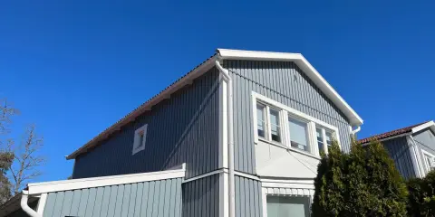 Blå himmel över ett gråblått trähus. Snart dags att måla fasad för att skydda det mot väder och vind.