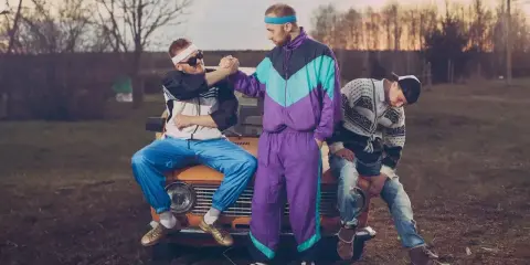Trzech kumpli w dresach z lat 90. szykuje się na imprezę. Jeden siedzi na masce, drugi podaje mu rękę, trzeci odpoczywa obok.