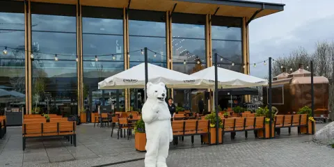Pluszowy miś wita gości przed nowoczesną karczmą na wierchu. W tle ogródek z parasolami i miedziane kadzie.