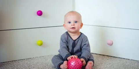 7 miesięczne dziecko z niebieskimi oczami siedzi na dywanie, trzymając w rączkach różową piłeczkę.