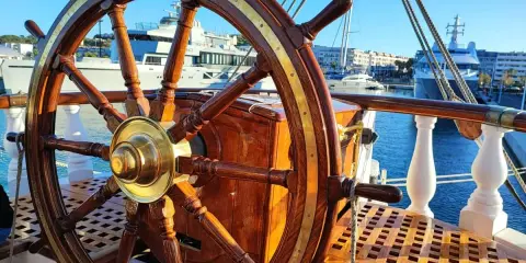 Roue de gouvernail en bois et laiton sur le pont d'un bateau. En arrière-plan, des yachts modernes et des bâtiments dans un port ensoleillé.