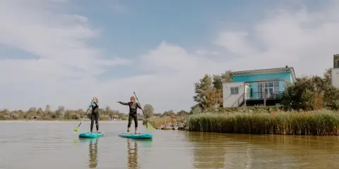 Zwei Frauen auf SUP-Boards am Makkum Strand, mit einem modernen Haus im Hintergrund.