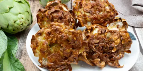 Carciofi alla giudia dorati e croccanti su un piatto bianco, con carciofi freschi sullo sfondo.