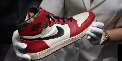 Ein Paar rote und weiße Air Jordan 1 Sneaker, die als die teuersten Schuhe der Welt gelten, werden von Händen in weißen Handschuhen gehalten.
