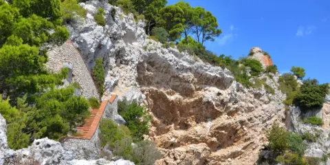Scogliera rocciosa con una scalinata in mattoni che sale verso la cima, dove si intravede un rudere. Si immagina il **salto di Tiberio** da questo luogo suggestivo.