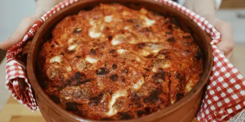 Un invitante pasta al forno appena sfornata, con crosta dorata e ripieno filante, tenuta con un canovaccio a quadretti rossi.
