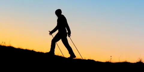 Sylwetka osoby uprawiającej nordic walking na tle zachodzącego słońca. Czy wolisz nordic walking rano czy wieczorem?