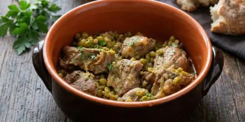 Un saporito agnello con piselli, cotto lentamente in una ciotola di terracotta, guarnito con prezzemolo fresco.