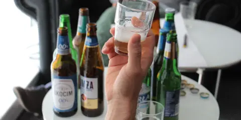 Ręka trzyma kieliszek z piwem bezalkoholowym. W tle kilka butelek piwa, idealne na przerwę w pracy.