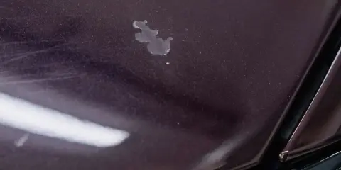 Na ciemnym lakierze widoczne przetarcie do bazy, z widocznymi rysami i plamą.