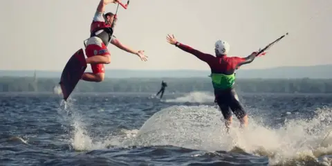 Zwei Kitesurfer springen und reichen sich die Hände auf dem Steinhuder Meer. Ein weiterer Surfer im Hintergrund.