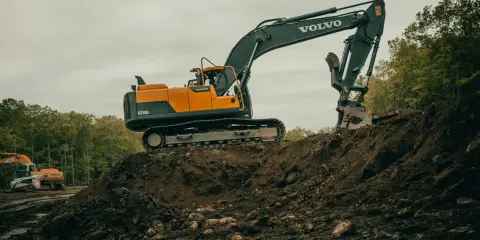 Pomarańczowo-czarna koparka Volvo na placu budowy, z innym mniejszym sprzętem w tle.
