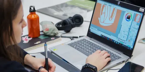 Studentka pracuje nad projektem w **studiach graficznych Warszawa**. Na ekranie laptopa widać program do grafiki, a obok słuchawki i butelkę z wodą.