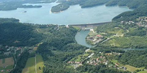 Widok z lotu ptaka na zapora wodna w Myczkowcach, otoczona zielonymi wzgórzami i rozległym jeziorem.