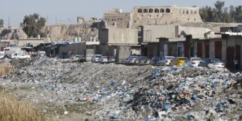 Müllberge säumen die Straßen von Kirkuk, Irak. Autos parken hinter den Geschäften, während im Hintergrund alte Gebäude auf einem Hügel stehen.