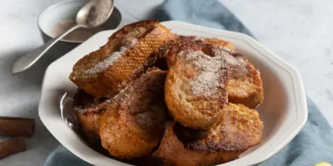 Un plato blanco lleno de torrijas españolas doradas y espolvoreadas con azúcar y canela.