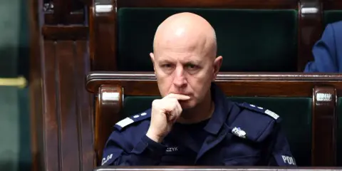 Łysy mężczyzna w mundurze policji siedzi w drewnianej ławie, zamyślony.