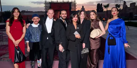 A group poses at a fashion event in Arabia. The backdrop features a large screen with "FASHION TRUST ARABIA" visible.