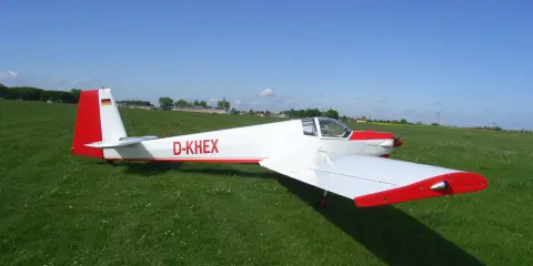 Ein weißer Motorsegler Falke mit roten Akzenten steht auf einer grünen Wiese.