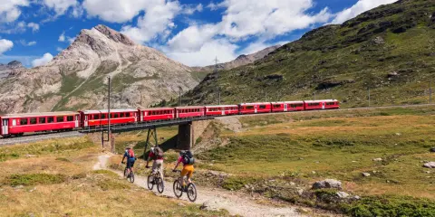 Treno rosso attraversa un ponte su un paesaggio alpino mozzafiato. Tre ciclisti pedalano su un sentiero sottostante, godendosi la vista della ferrovia del Bernina.