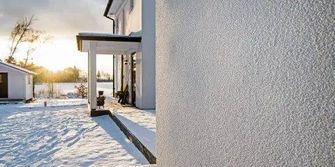 Białe, chropowate tynki na ścianach domu w zimowy, słoneczny dzień. Śnieg pokrywa podwórko i otoczenie.