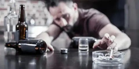 Mężczyzna z papierosem w popielniczce, butelki i kieliszek na stole. Nadmierne spożycie alkoholu może pogorszyć hemoroidy.