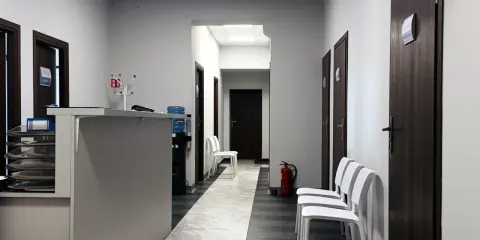 Korytarz kliniki medycznej z recepcją, drzwiami i białymi krzesłami.