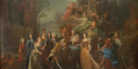Max Emanuel Herzog in Bayern, umgeben von Adeligen und allegorischen Figuren, in einer prunkvollen Szene vor einem Schloss.
