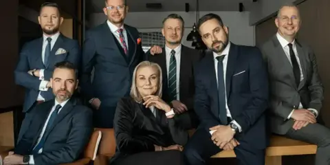 Grupa siedmiu profesjonalistów w garniturach, pozujących do zdjęcia w biurze.