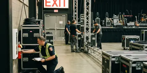 Pracownik sprawdza gaśnicę przy znaku "EMERGENCY EXIT". W tle widać scenę koncertową i sprzęt. To miejsce zbiórki do ewakuacji.