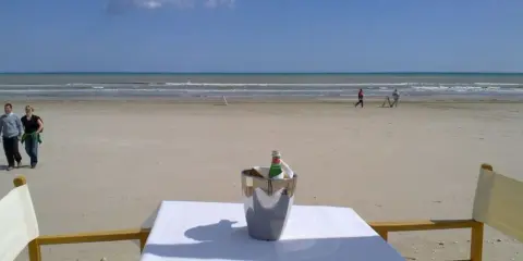 Coppia passeggia sulla spiaggia di Milano Marittima, con un secchiello del ghiaccio e una bottiglia su un tavolo in primo piano.