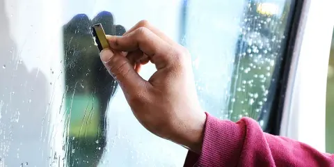 Dłoń z użyciem skrobaka usuwa resztki kleju z szyby. Dowiedz się, czym zmyć klej z szyby, by uzyskać idealną czystość.