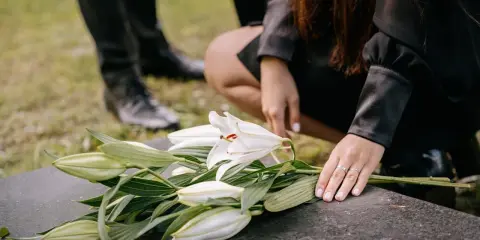 Dłoń kładzie białe lilie na kamieniu nagrobnym. W tle widać nogi w czarnych butach. To symboliczny gest, który może nastąpić w dowolnym momencie po śmierci, nie ma ustalonego terminu, ile dni po śmierci jest pogrzeb.