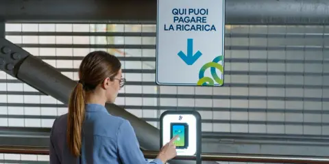 Donna interagisce con un terminale per pagare la ricarica dell'auto elettrica. Il cartello indica "Qui puoi pagare la ricarica", suggerendo il costo ricarica auto elettrica.