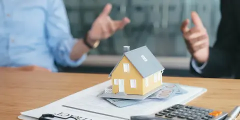 Rozmowa o umowie najmu krótkoterminowego a VAT. Na stole model domu, pieniądze, kalkulator i dokumenty.