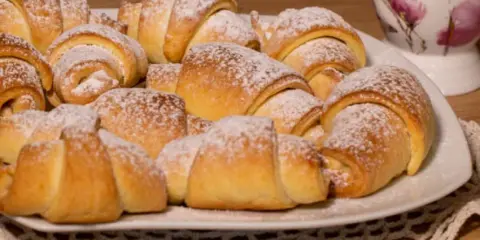 Talerz pełen puszystych, posypanych cukrem pudrem rogalików krucho drożdżowych. Idealne na śniadanie lub deser.